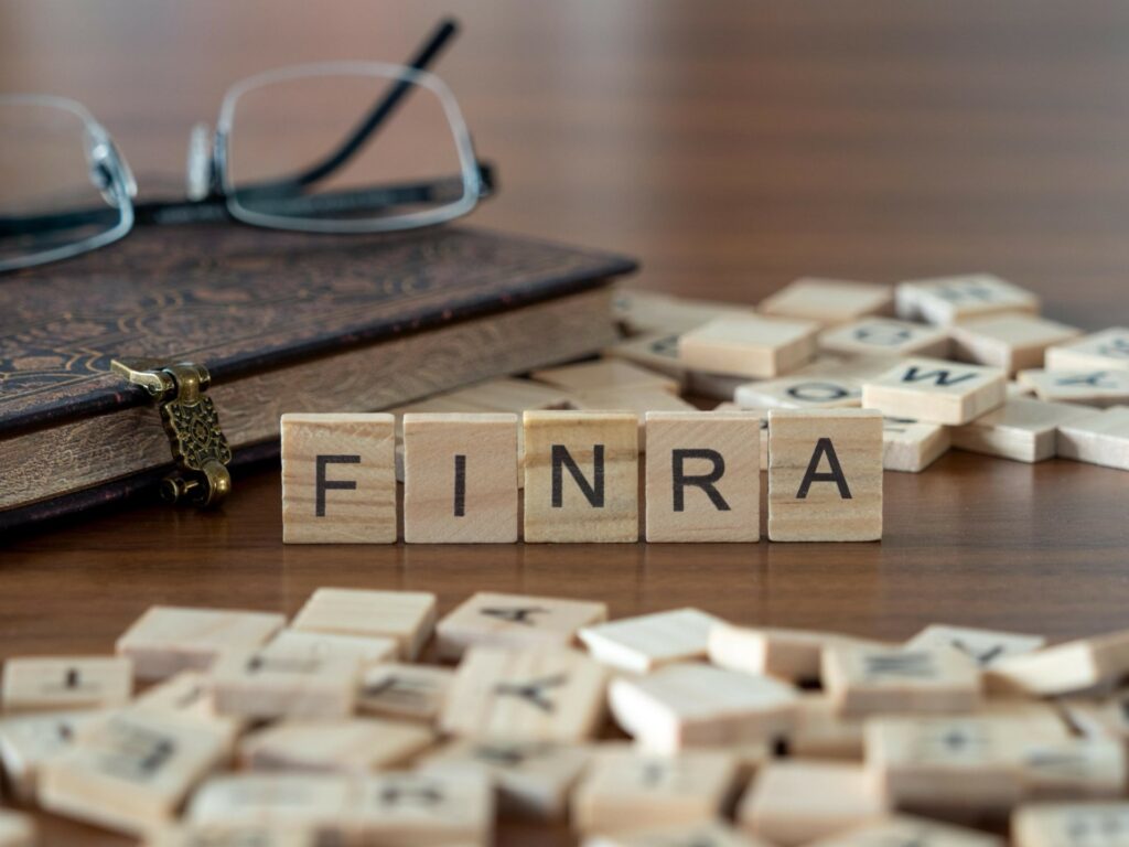 the acronym finra for financial industry regulatory authority in wooden letter tiles on a wooden table with glasses and a book