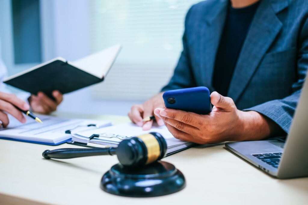 One person holding a pen and notebook while the other holds a phone next to paperwork, gavel and laptop